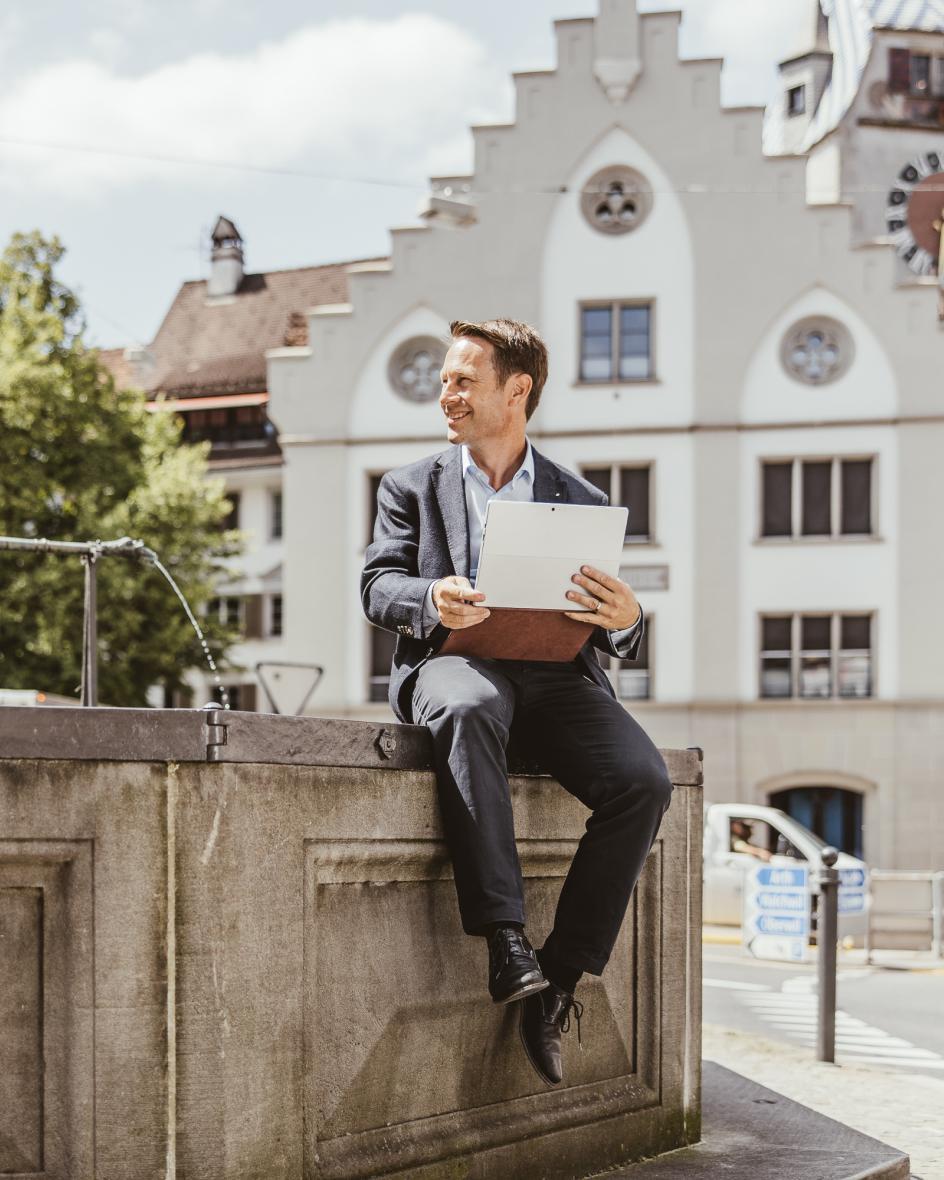 Ein Geschäftsmann sitzt auf einem Brunnen in der Altstadt Zug. Im Hintergrund ist der Zytturm zu sehen.